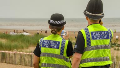Two police officers wearing high‑visibility ‘Heddlu Police’ vests looking out over a beach with people in the distance.