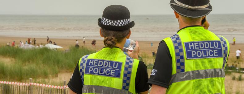 Two police officers wearing high‑visibility ‘Heddlu Police’ vests looking out over a beach with people in the distance.