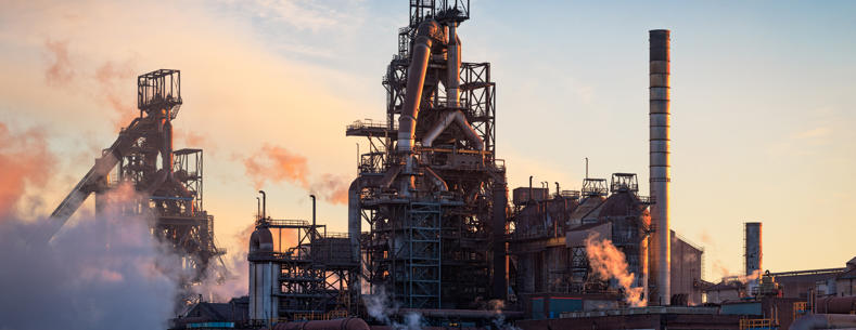 Equipment and piping of the Tata steel plant in Port Talbot with steam emissions in the foreground and sunset in the background.