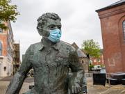A bronze statue of Dylan Thomas seated outdoors in a paved courtyard, wearing a face mask, with brick buildings and trees in the background.