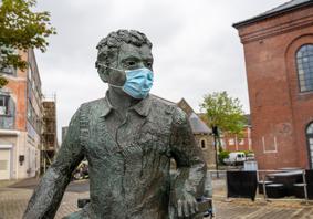 A bronze statue of Dylan Thomas seated outdoors in a paved courtyard, wearing a face mask, with brick buildings and trees in the background.