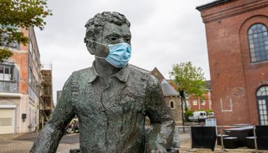 A bronze statue of Dylan Thomas seated outdoors in a paved courtyard, wearing a face mask, with brick buildings and trees in the background.
