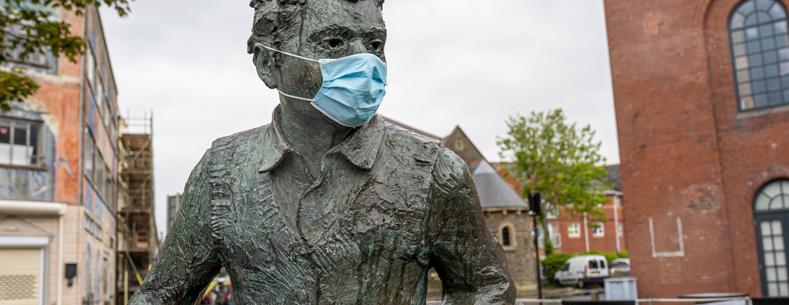 A bronze statue of Dylan Thomas seated outdoors in a paved courtyard, wearing a face mask, with brick buildings and trees in the background.
