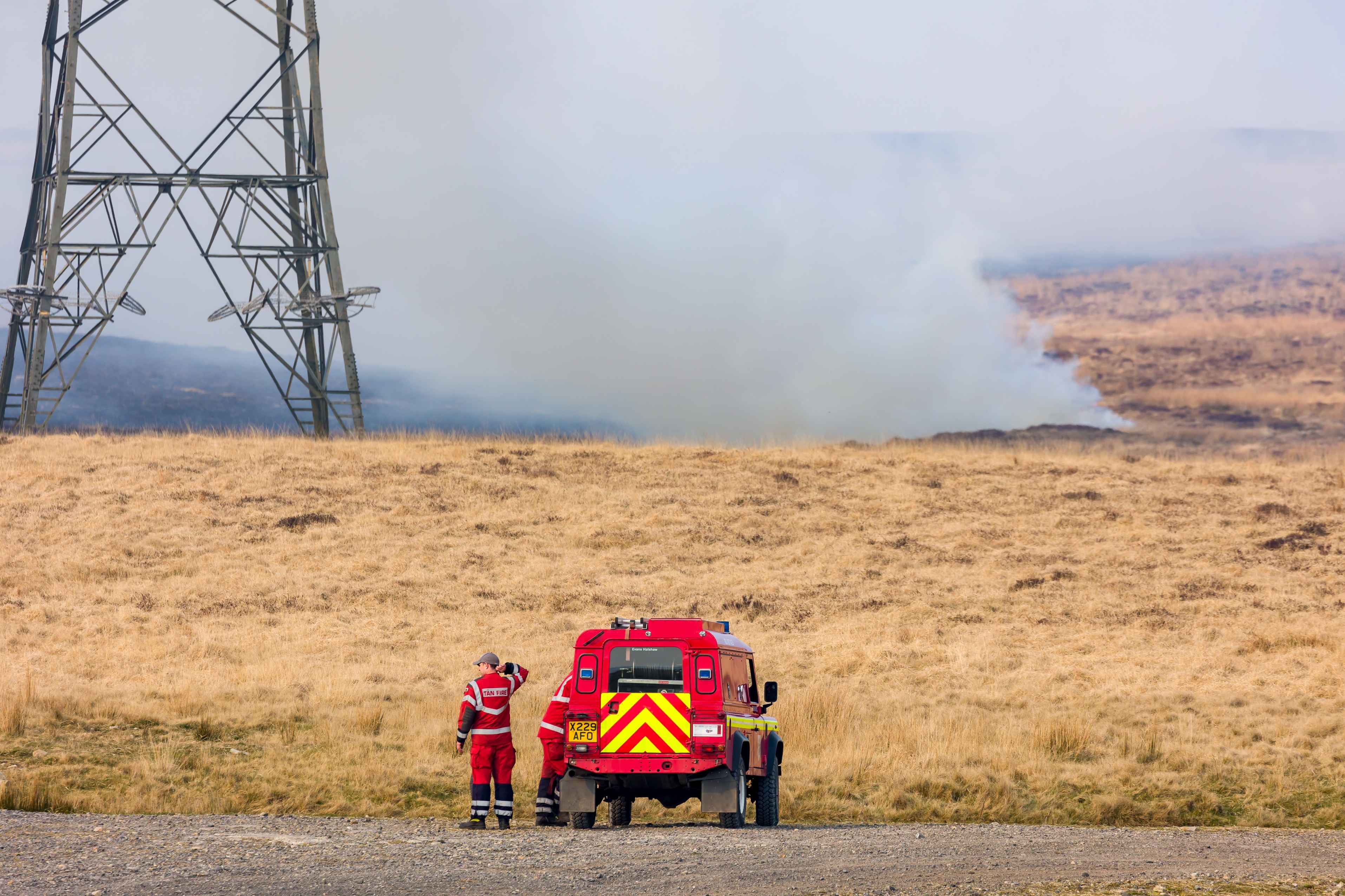 A landscape photograph of a fire engine in front of a moorland grass fire, with smoke rising up towards a pylon in the background.
