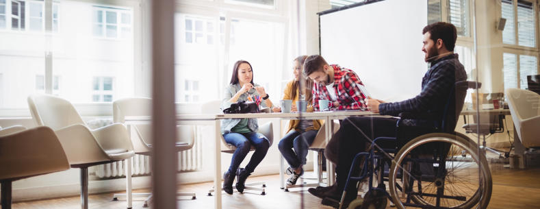 An image of a workplace with 4 people sat around a table working, including one wheelchair user