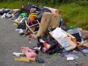 A pile of fly-tipped rubbish dumped on a road, including furniture, black bin bags, a sofa, cushions, toys, and household items scattered on the ground beside grass.
