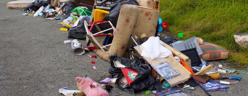 A pile of fly-tipped rubbish dumped on a road, including furniture, black bin bags, a sofa, cushions, toys, and household items scattered on the ground beside grass.