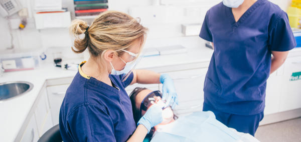 A dentist’s room with a patient wearing protective glasses lying back in the dentist chair. A dental worker wearing a mask, gloves and protective glasses is looking in the patient’s mouth. Another dental worker, also wearing a mask, looks on.