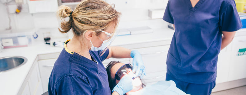 A dentist’s room with a patient wearing protective glasses lying back in the dentist chair. A dental worker wearing a mask, gloves and protective glasses is looking in the patient’s mouth. Another dental worker, also wearing a mask, looks on.