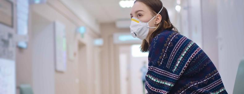 A young woman sitting alone in a hospital corridor with a face mask on waiting for someone to come.