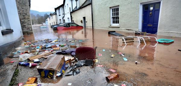 This is a picture of a street flooded by muddy water with floating rubbish and domestic items in the foreground. In the background are vehicles, most of which are submerged, one is  There is one floating car.