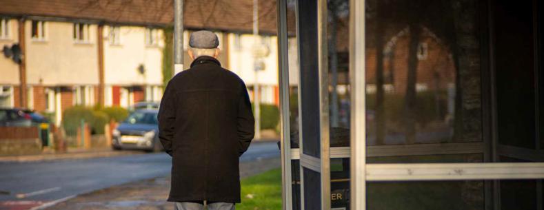 Older man standing at bus stop facing away from the camera.